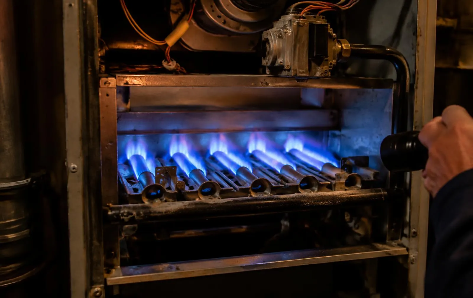 Gas burner flames lit inside an open furnace cabinet, lit by a technician's flashlight